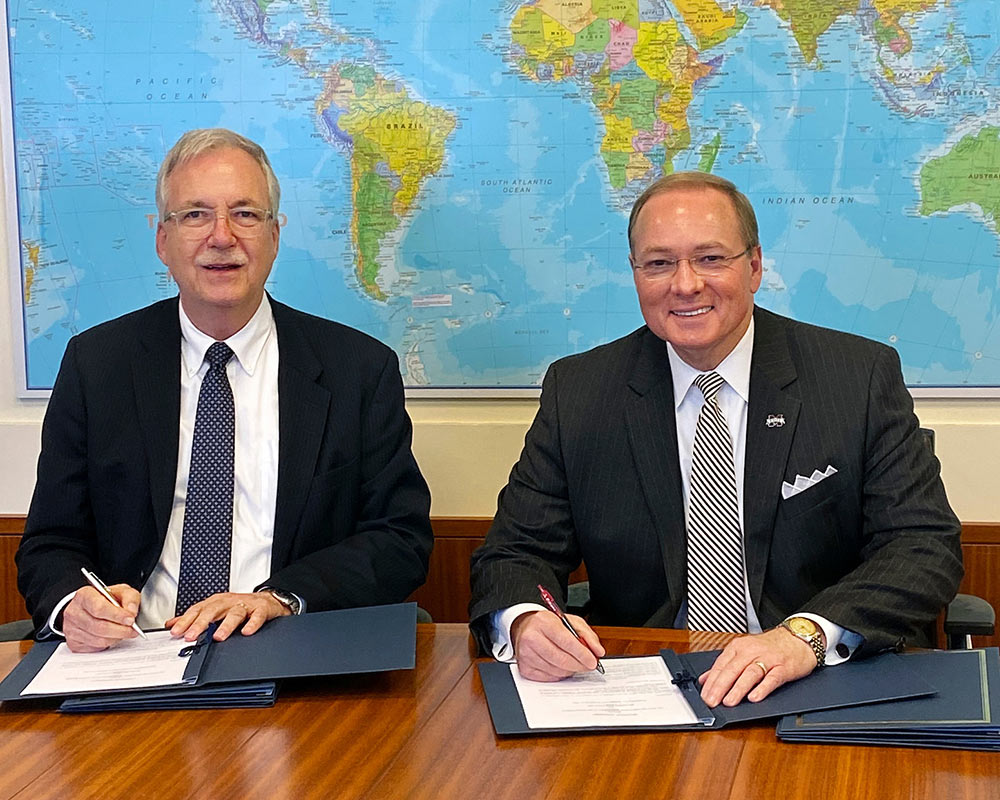 Two men seated at a table holding pens to sign documents. A world map is on the background wall.